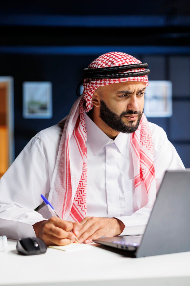 An Arab businessman, engrossed in work, efficiently browses digital content on a laptop at his office desk. Taking notes, he conducts online research, showcasing his professionalism and dedication.